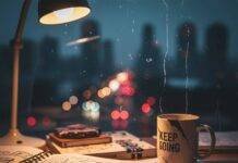 Why Mental Health Education is Critical for Our Future—A Deep Dive A blurry, photorealistic image of a messy desk with a doodle-filled notebook, a chipped "Keep Going" mug, and a city skyline visible through a streaky window.