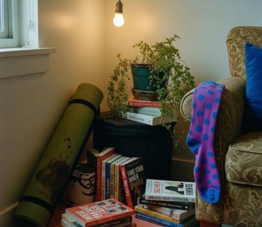 Holistic Health and Wellness: 10 Steps to Start Living Your Best Life A dimly lit corner of a cluttered apartment features a rolled-up, coffee-stained yoga mat, stacks of self-help books, a drooping fern, and a bright purple polka-dot sock hanging from an armchair.